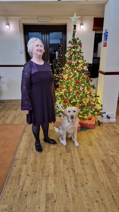 Secretary Sue in a glitzy plum coloured dress stood with guide dog robin in front of a decorated Christmas tree complete with sparkly lights.